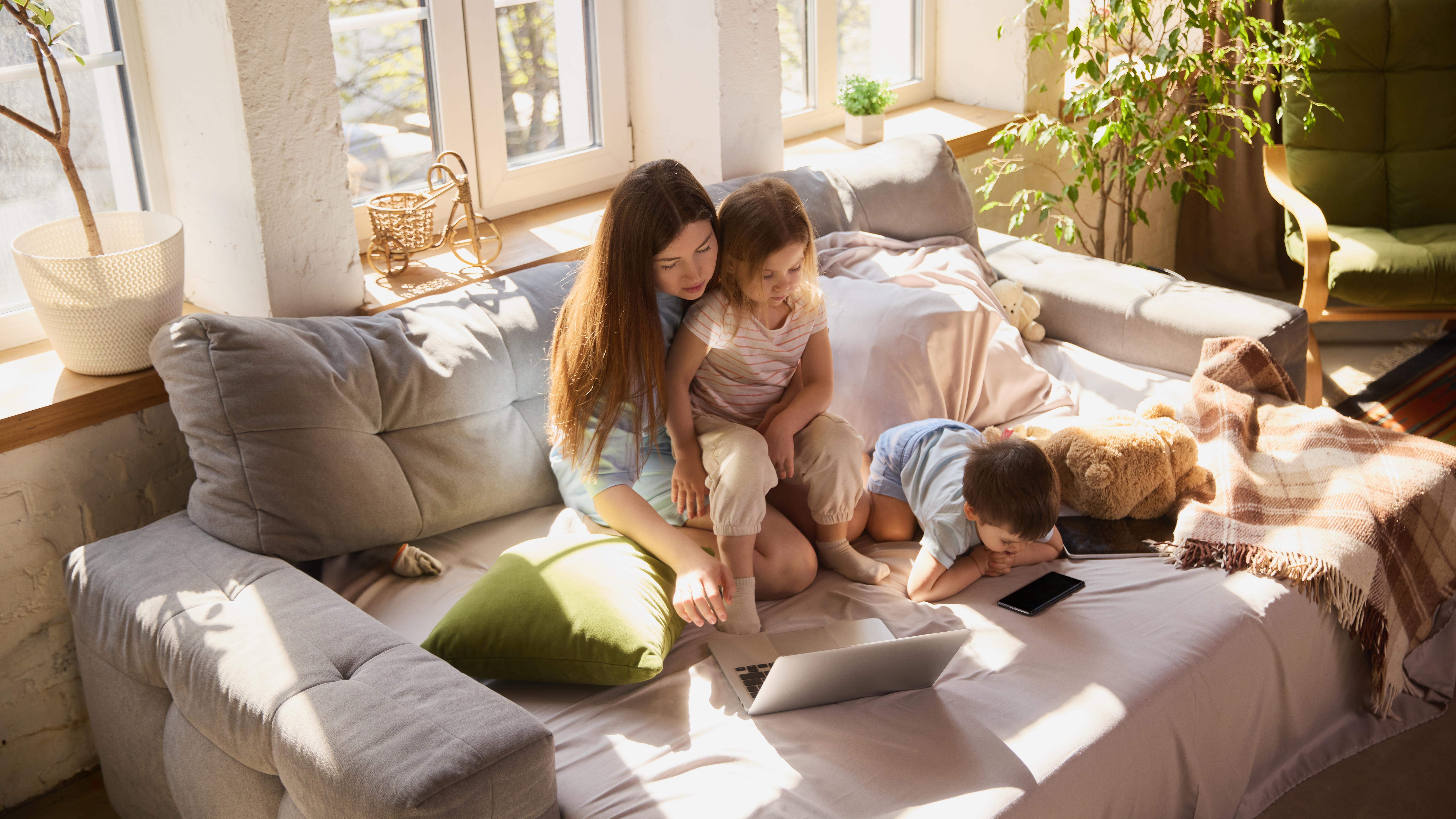 Mother with children using laptop together on sofa in bright living room. Concept of online homework, home education, parental guidance, screen time management and daily routine.