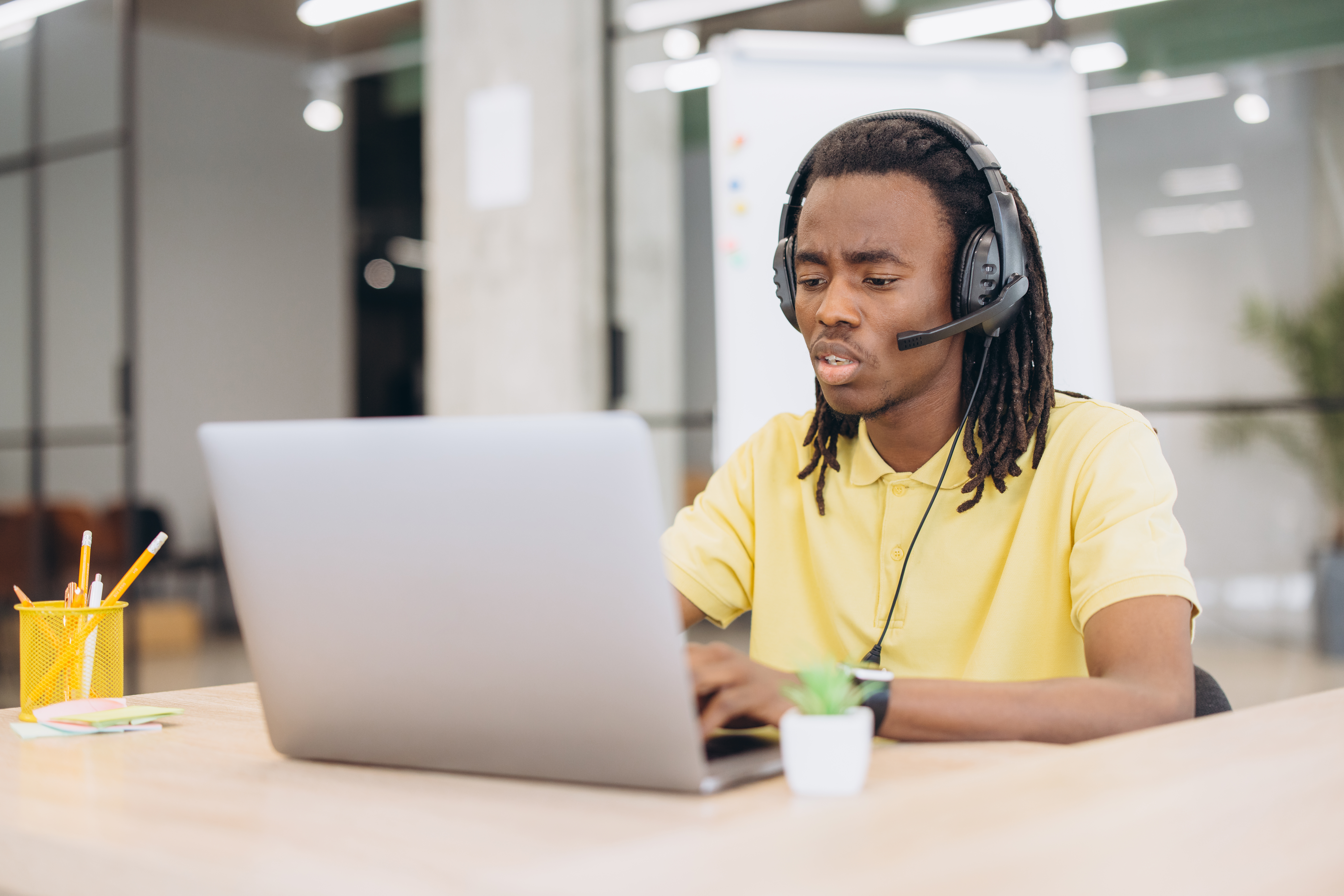 Black man wearing a headset and yellow polo, typing on a laptop, concentrating during an online conference call or remote work